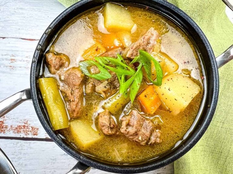 A bowl of beef stew with chunks of beef, potatoes, carrots, and garnished with sliced green onions.
