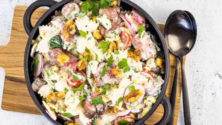 A bowl of pasta salad with steak slices, cherry tomatoes, radishes, walnuts, greens, and creamy dressing, served on a wooden board with a black spoon and fork.
