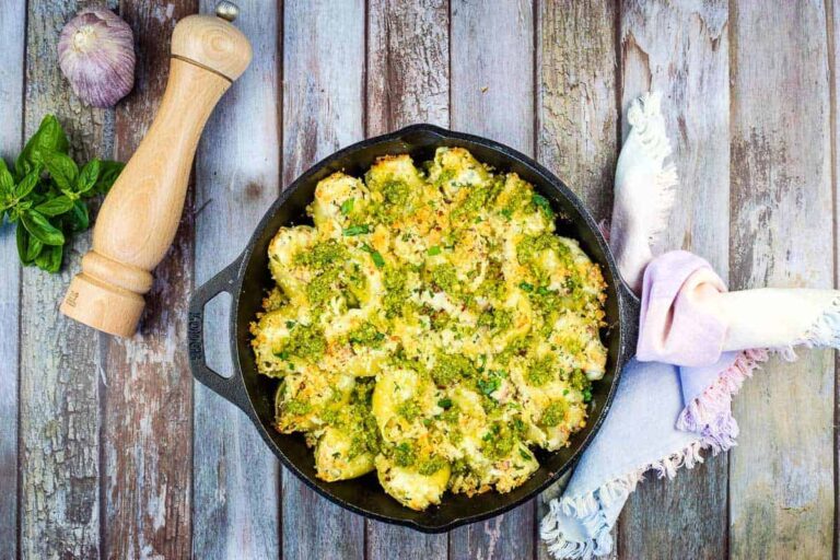 A cast iron skillet filled with baked stuffed pasta shells topped with green sauce and breadcrumbs, next to a pepper grinder, garlic, basil, and a fringed napkin on a rustic wooden surface.