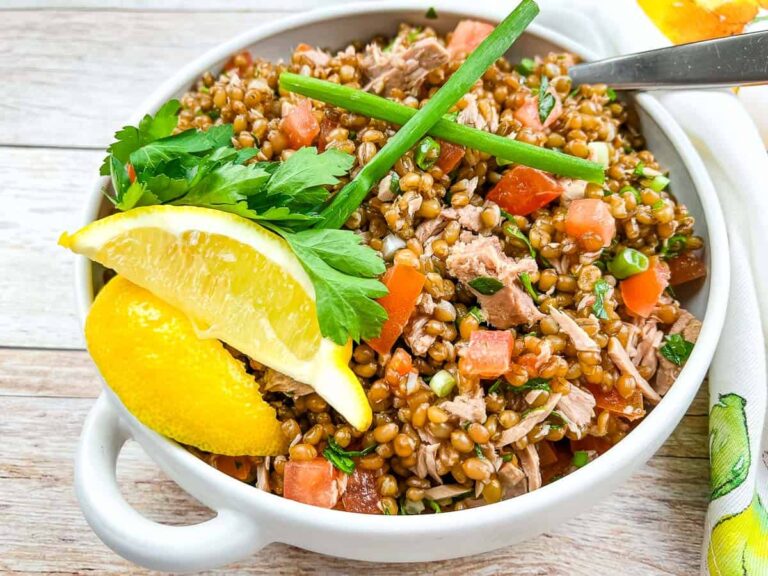 A bowl of grain salad with chopped tomatoes, herbs, shredded meat, and two lemon wedges, garnished with parsley and green onion on a wooden surface.