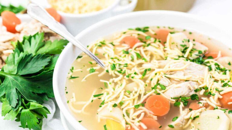 A bowl of chicken noodle soup with carrots, herbs, and shredded chicken, with a spoon in the bowl and fresh parsley beside it.