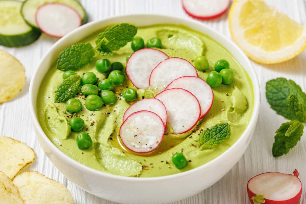 A bowl of Green Pea Hummus with Mint, topped with sliced radishes, green peas, mint leaves, and black pepper, with lemon, cucumber, chips, and radishes on the side.