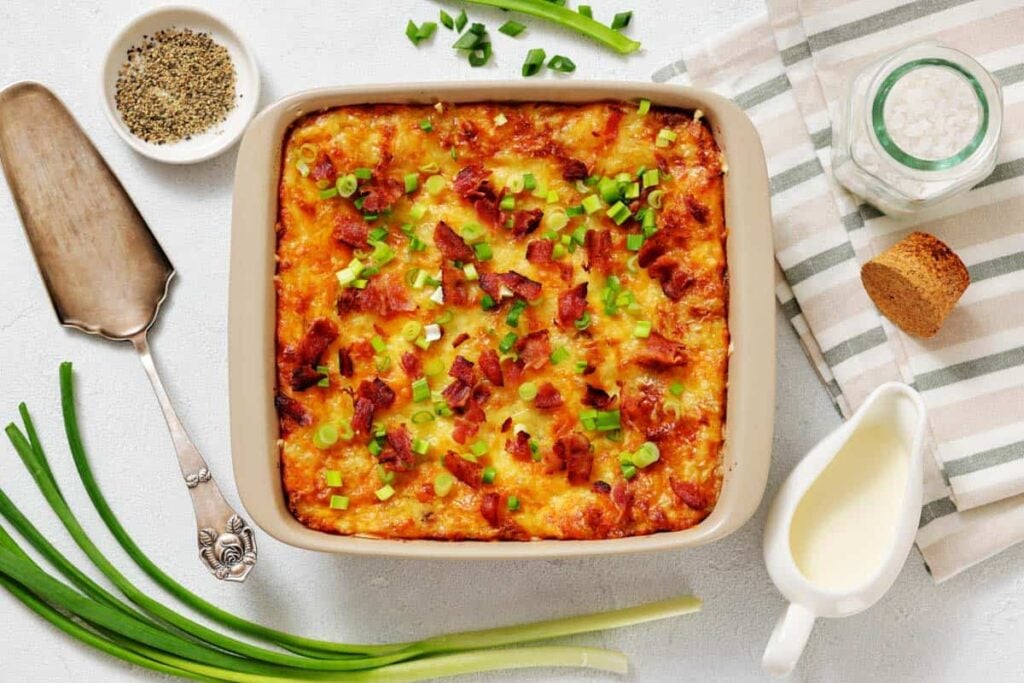 A baked casserole topped with chopped green onions and bacon bits, surrounded by green onions, a serving spatula, seasoning, and a striped napkin.