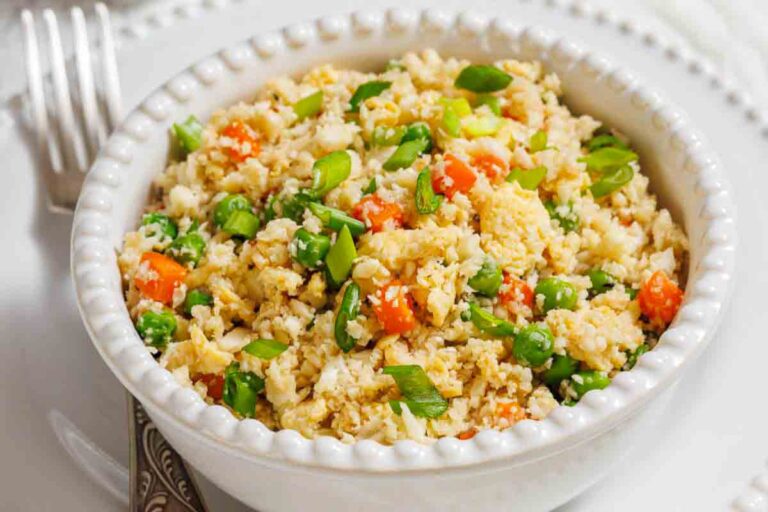 A white bowl filled with cauliflower fried rice, mixed with green peas, diced carrots, and chopped green onions, placed on a white plate with a fork beside it.