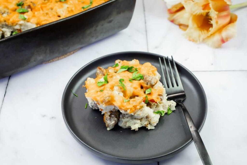 A serving of cheesy casserole with ground meat and herbs on a black plate with a fork, next to a casserole dish and pasta shells on a white surface.