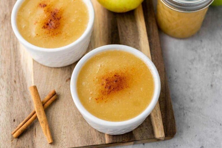 Two white ramekins filled with applesauce topped with cinnamon, placed on a wooden board next to cinnamon sticks and a jar of applesauce.