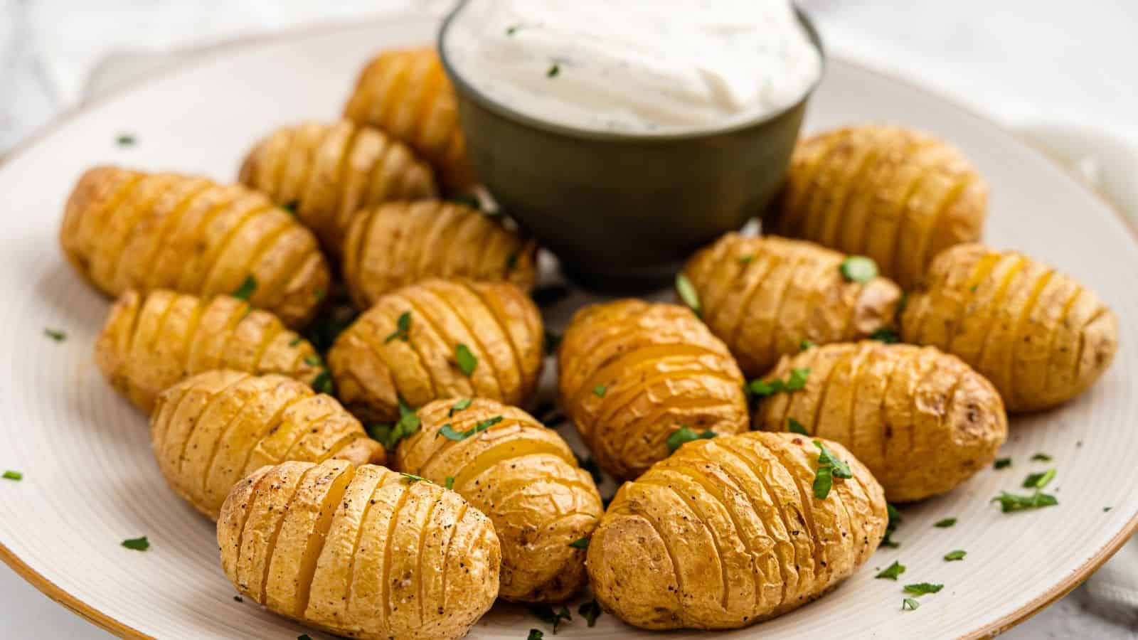 A plate of sliced, roasted baby potatoes is garnished with herbs. A small bowl of creamy sauce is placed in the center.