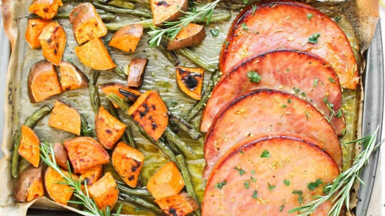 Sheet pan with sliced glazed ham, roasted sweet potato chunks, and green beans, garnished with fresh rosemary.