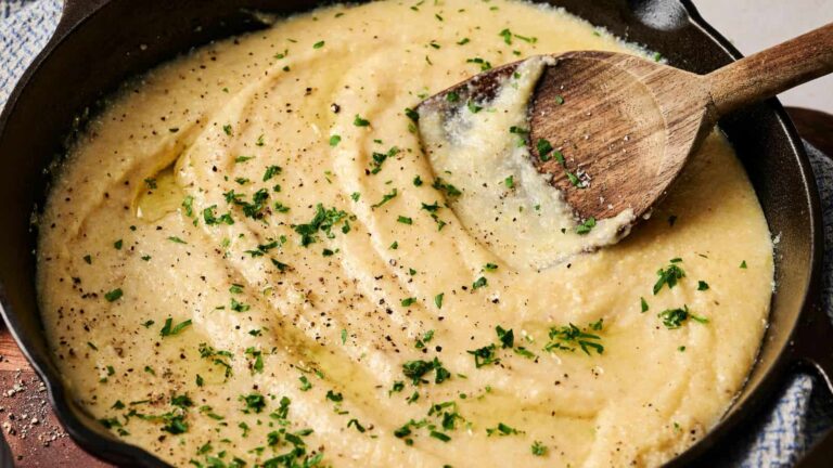 Creamy polenta topped with chopped herbs and cracked black pepper in a cast iron skillet, stirred with a wooden spoon.
