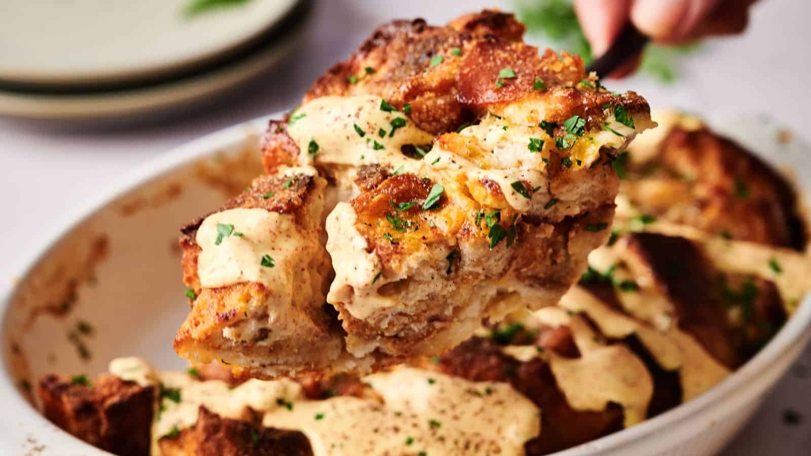 A serving of bread pudding with a creamy sauce and chopped herbs is lifted from a baking dish.
