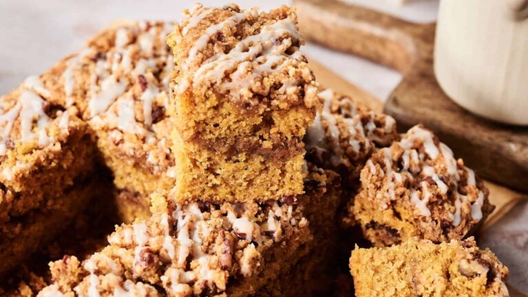 A stack of crumb-topped pumpkin coffee cake squares with white icing drizzle, arranged on a wooden surface.
