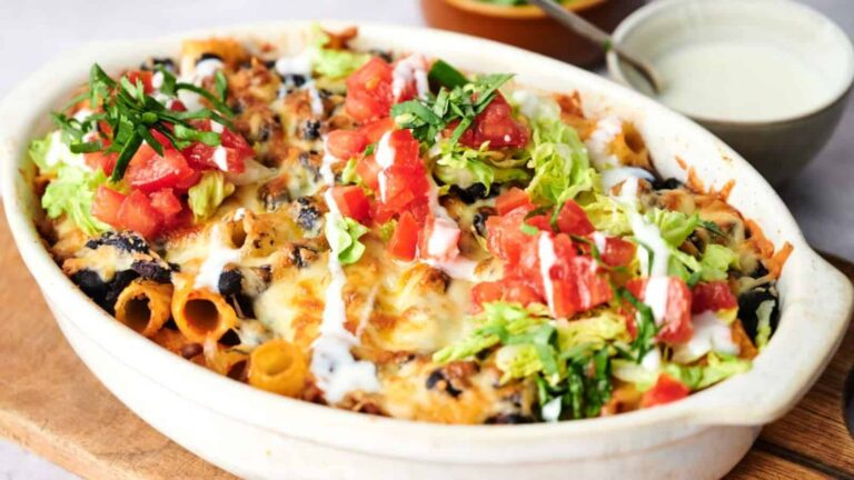 A baked casserole topped with melted cheese, lettuce, diced tomatoes, herbs, and drizzled with white sauce, served in a white oval dish on a wooden board.