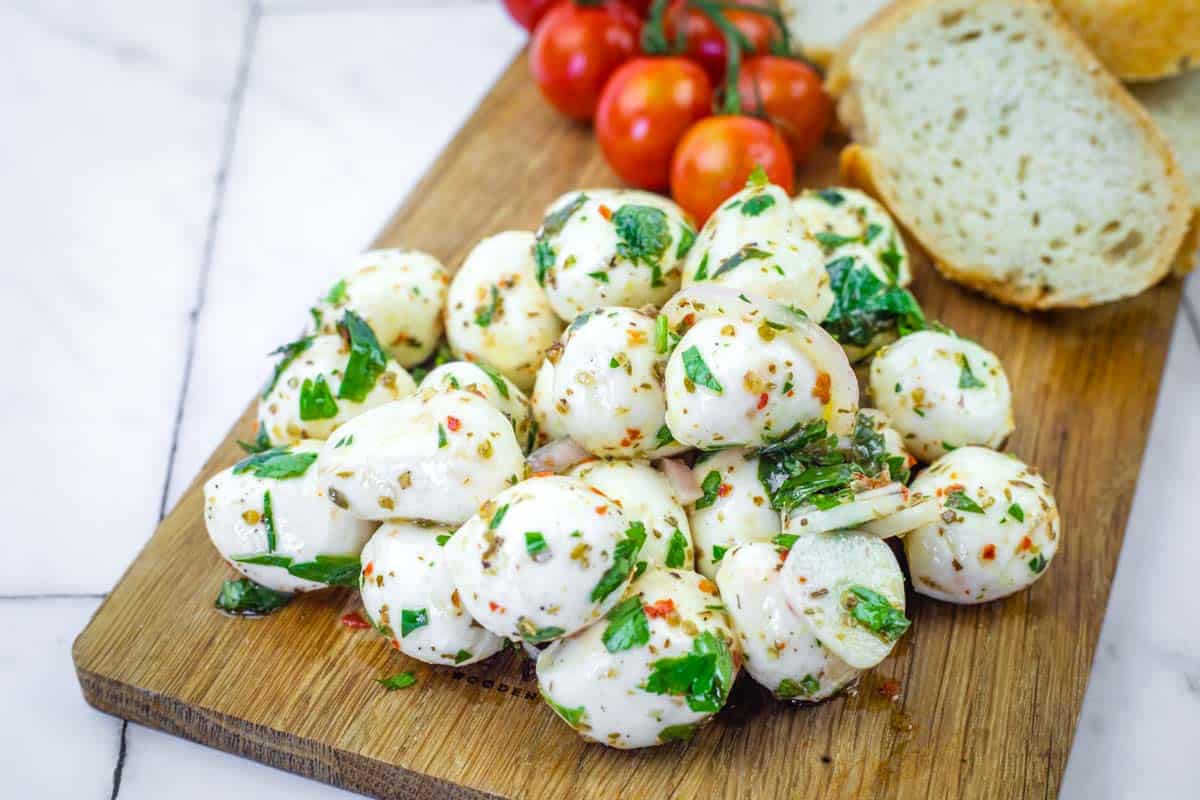A pile of herb-marinated mozzarella balls with herbs on a wooden board, next to sliced bread and a bunch of cherry tomatoes.