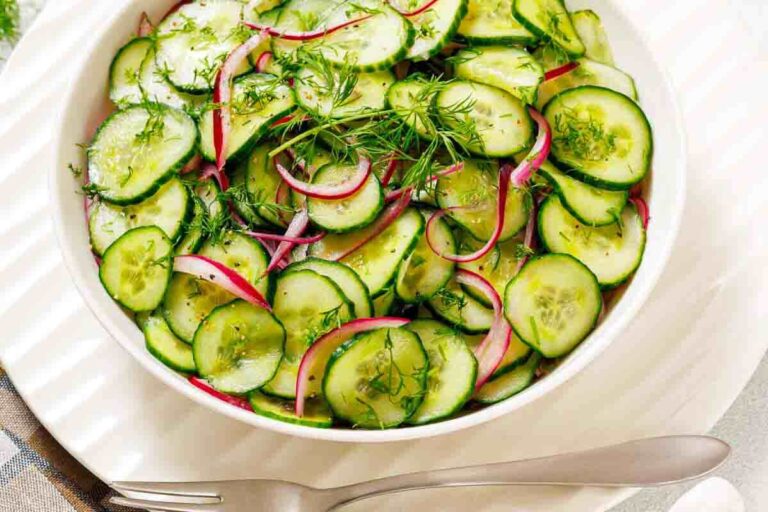 A bowl of cucumber salad with sliced red onions and fresh dill, placed on a white plate with a spoon and fork beside it.