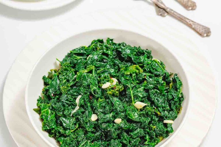 A white bowl filled with cooked kale and garlic slices, placed on a white plate with silverware in the background.