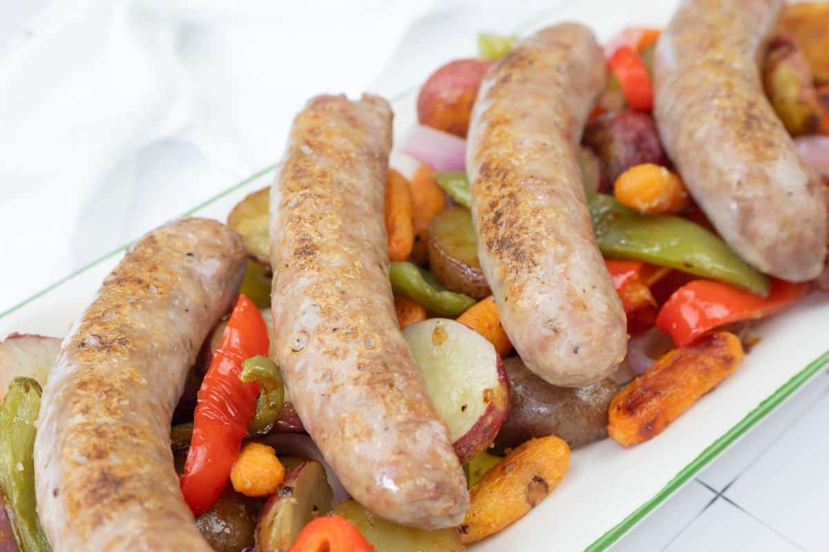 Four cooked sausages served on a platter with roasted potatoes, carrots, onions, green and red bell peppers.