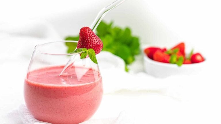 A glass of strawberry smoothie with a straw and a whole strawberry on the rim; a bowl of strawberries and green leaves in the background.