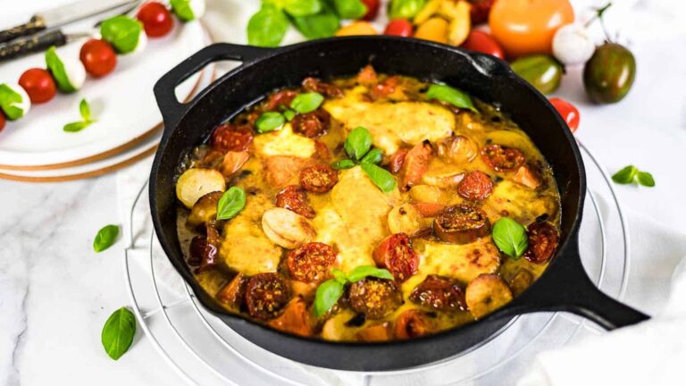 A cast iron skillet filled with baked chicken, tomatoes, sausage slices, and melted cheese, garnished with fresh basil leaves.
