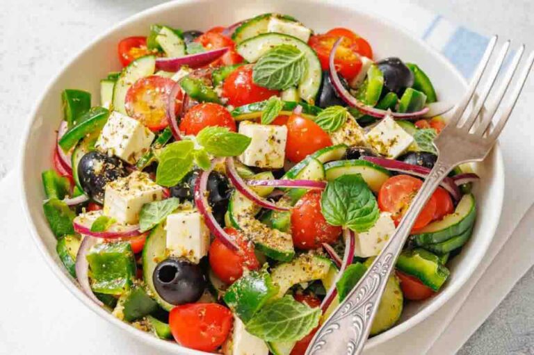 A bowl of Greek salad with cucumber, cherry tomatoes, feta cheese, black olives, red onion, green pepper, and mint leaves, served with a fork.