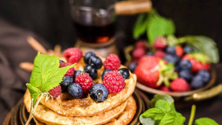 A stack of waffles topped with fresh raspberries, blueberries, and mint leaves, with a small bowl of mixed berries and a syrup container in the background.