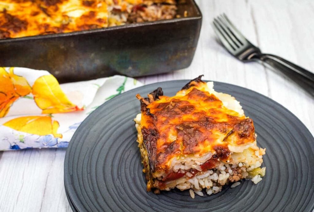 A slice of baked casserole with layers of rice, meat, and cheese on a black plate, with the rest of the casserole in a pan in the background.