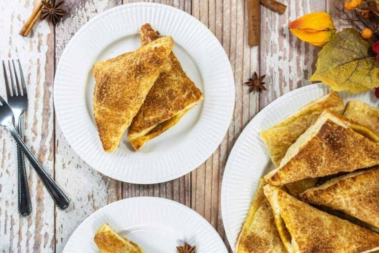 Plates with triangular pastries dusted with cinnamon sugar on a rustic wooden table, next to cutlery, spices, and autumn leaves.