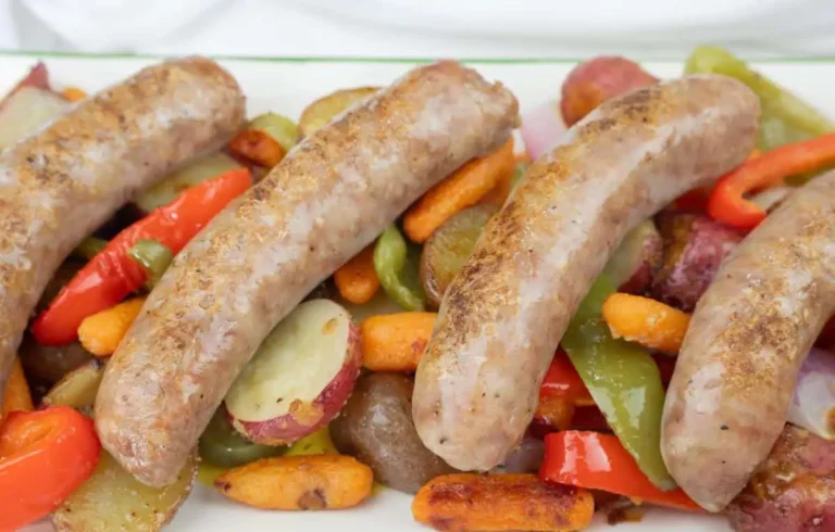 Four cooked sausages are served on a bed of roasted vegetables, including carrots, red potatoes, green bell peppers, and red bell peppers.