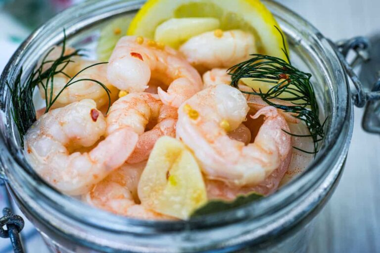 A glass jar filled with cooked shrimp, garnished with fresh dill, garlic slices, and a lemon wedge.