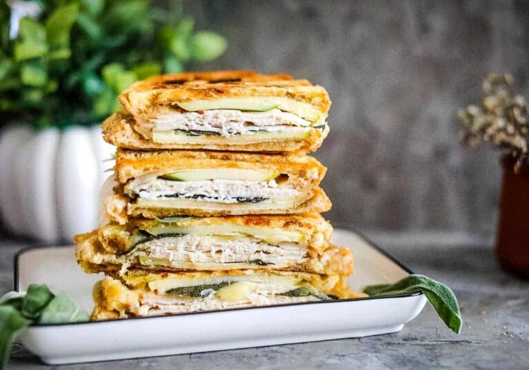 A stack of toasted sandwiches with visible layers of turkey, cheese, and sliced cucumbers on a white plate, with green plants in the background.