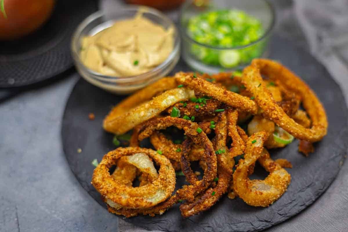 A pile of crispy onion rings garnished with chives, accompanied by small dishes of dipping sauces, are shown on a light wooden surface.