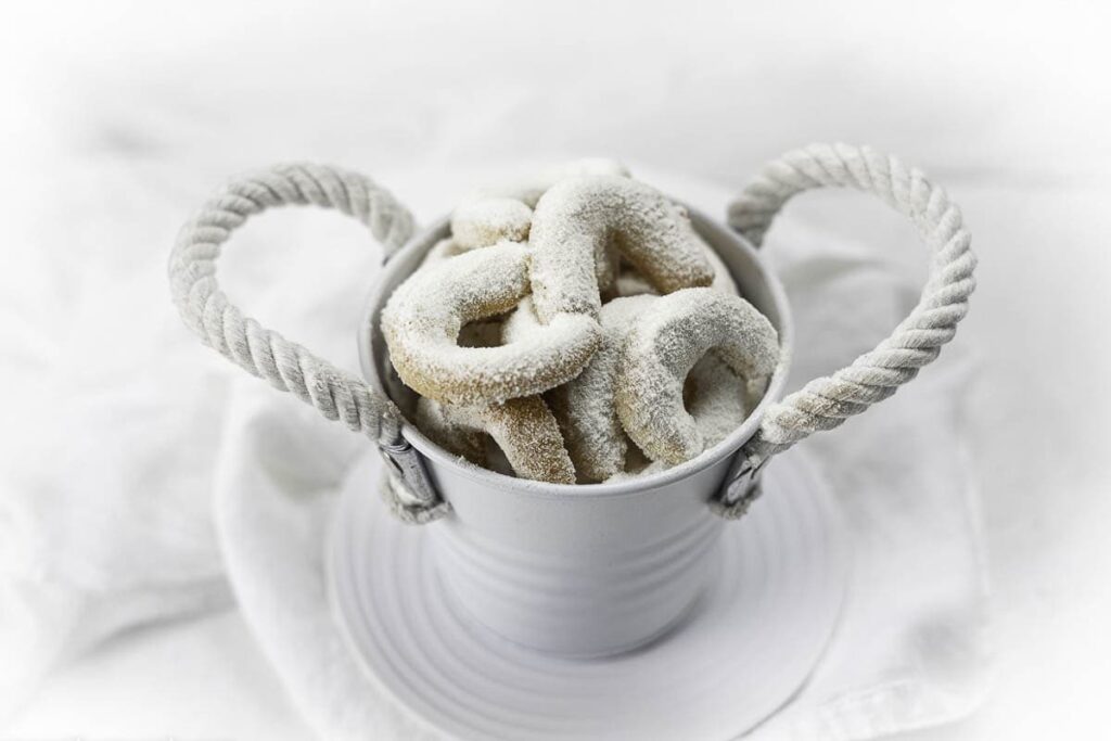 A small white bucket filled with powdered sugar-coated cookies, placed on a white plate against a white background.