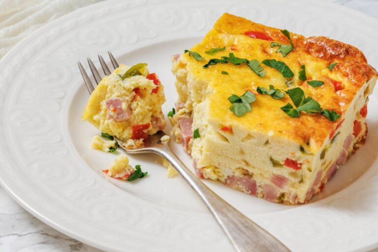 A slice of egg casserole with ham, red peppers, and herbs sits on a white plate with a fork holding a bite-sized piece.