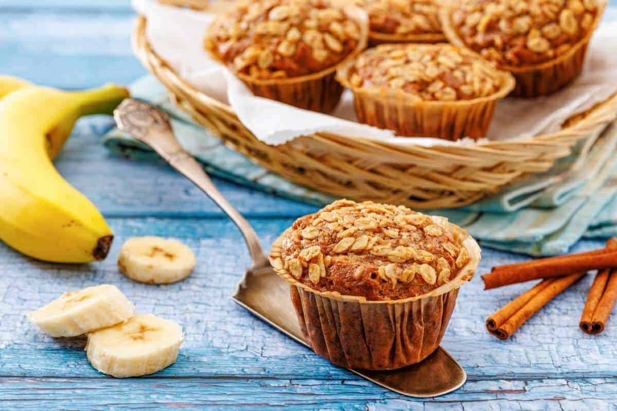 Banana oatmeal muffins on a metal spatula and in a basket, with sliced bananas and cinnamon sticks on a blue wooden surface.