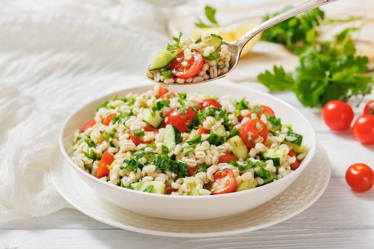 A bowl of barley salad with cherry tomatoes, cucumber, and parsley sits on a plate, with a spoonful of salad being held above the bowl.