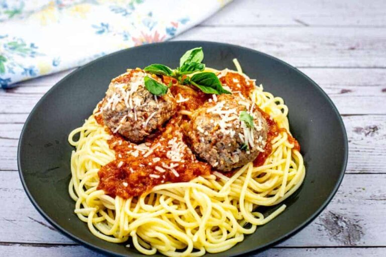 A black plate of spaghetti topped with tomato sauce, two meatballs, grated cheese, and a sprig of basil, placed on a light wooden surface.