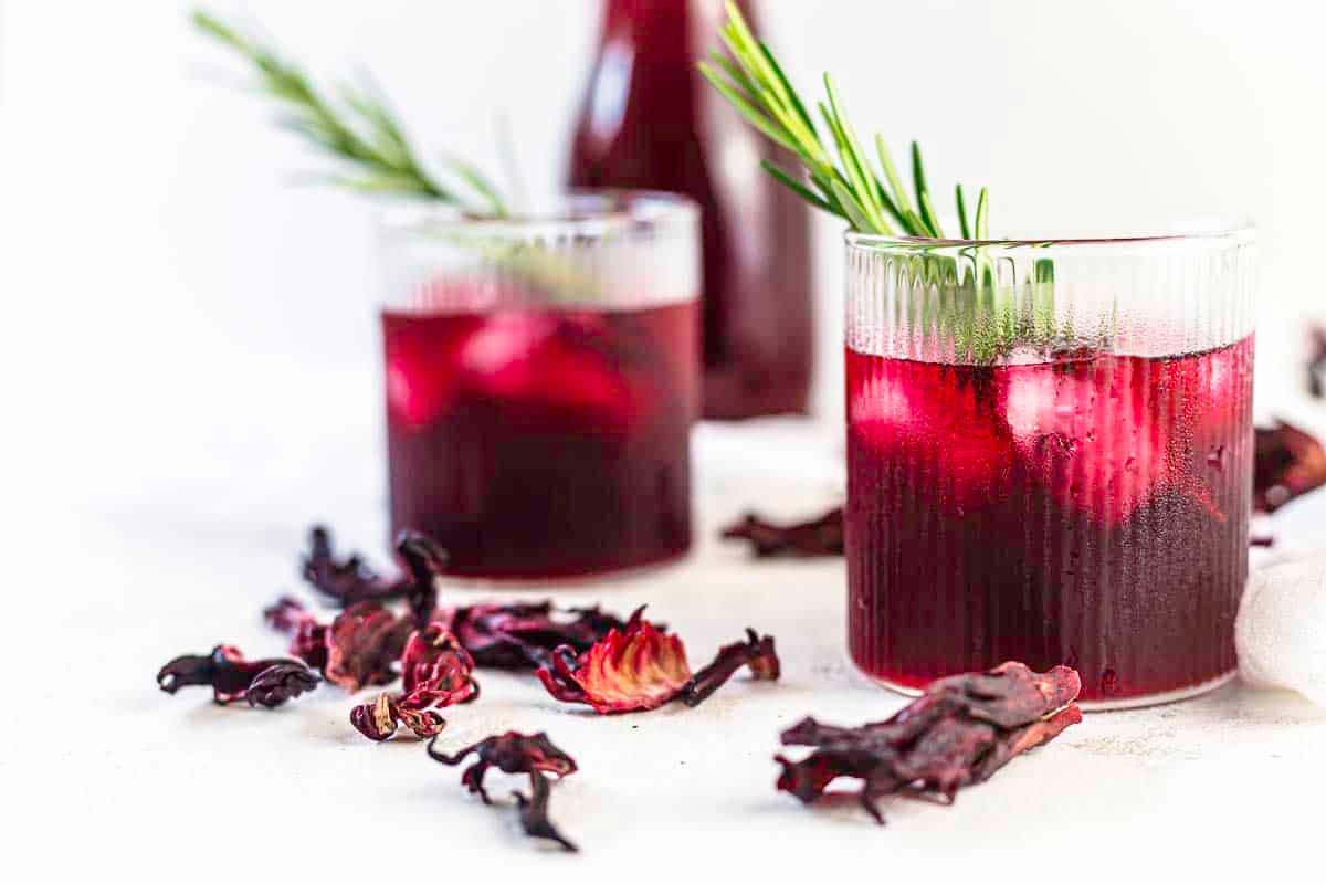 Two glasses of iced hibiscus tea garnished with rosemary sprigs, with dried hibiscus flowers scattered on a white surface. A bottle of the drink is in the background.