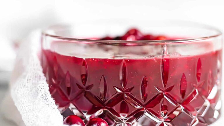 A close-up of a glass bowl filled with cranberry sauce, with a few whole cranberries visible on top and a textured cloth partially draped over the side.
