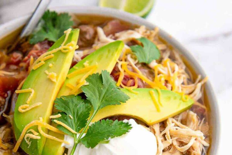 Bowl of shredded chicken soup topped with avocado slices, shredded cheese, cilantro, and a dollop of sour cream.