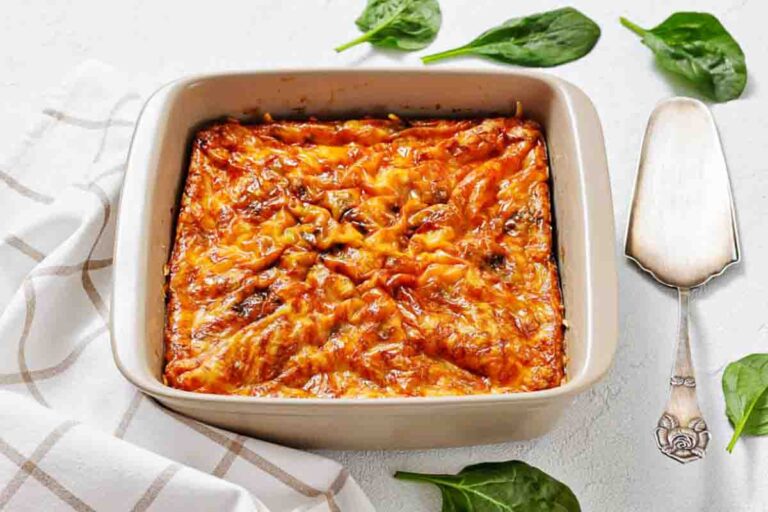A baked Sausage Egg Casserole in a square ceramic dish on a white surface, with spinach leaves and a serving utensil nearby.