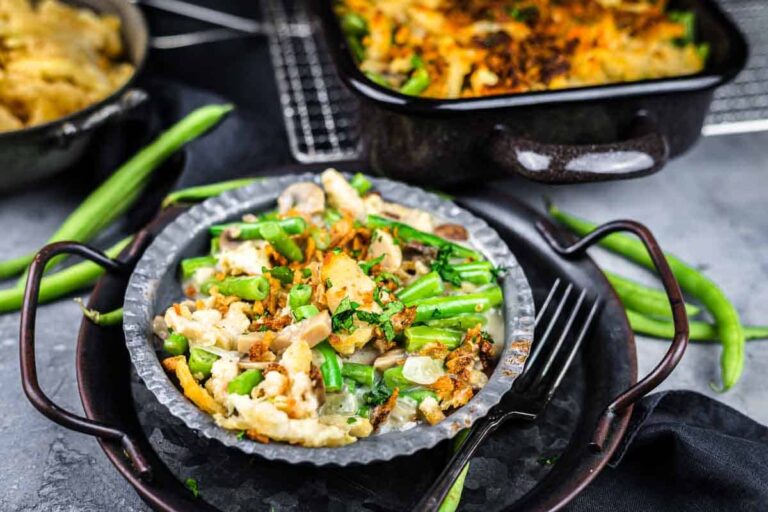 A plate of green bean casserole topped with crispy onions sits on a tray with a fork; the casserole dish is in the background with fresh green beans scattered nearby.
