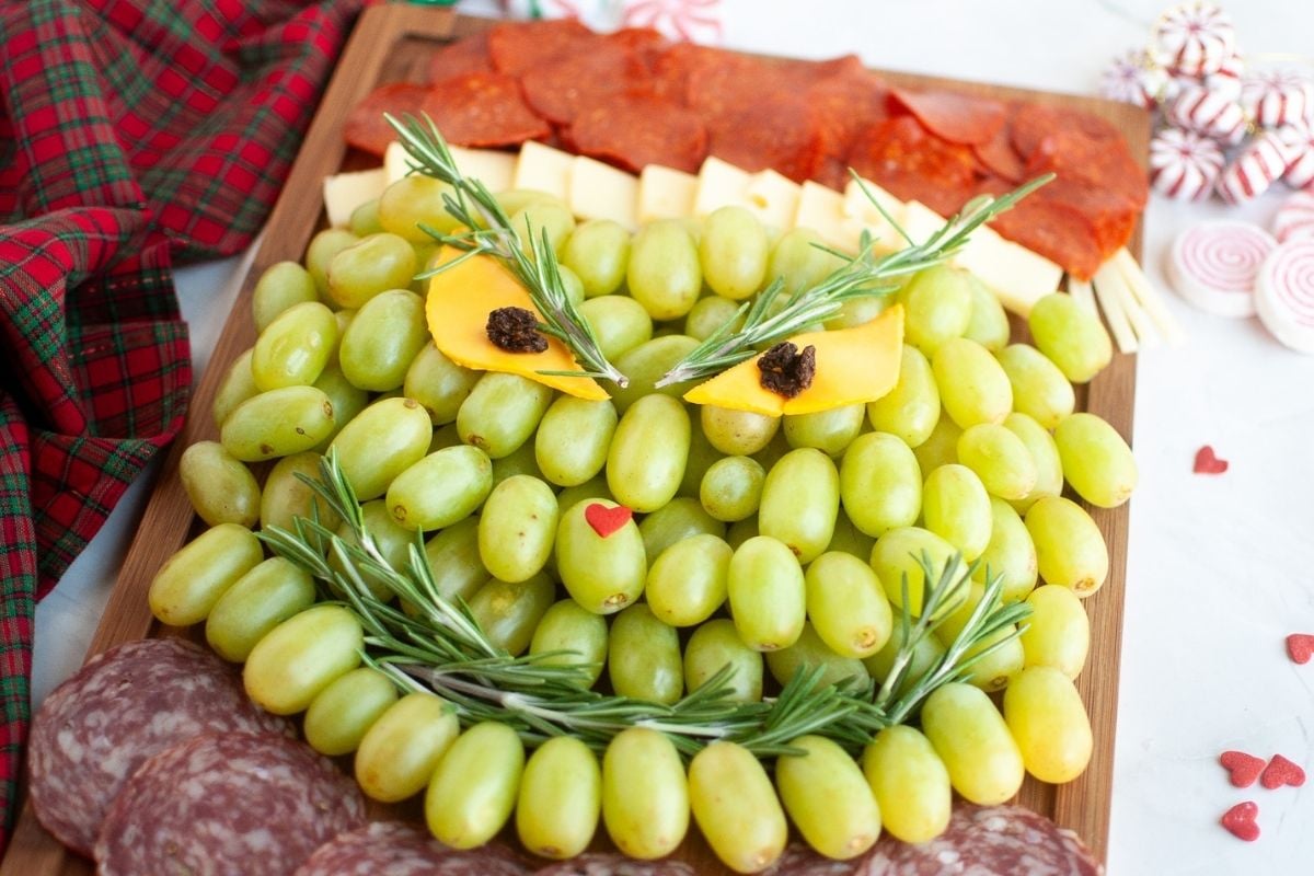 A charcuterie board arranged to resemble the Grinch's face, made with green grapes, cheese, meat slices, and garnished with rosemary.
