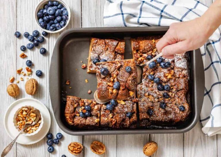 A hand serves a piece of baked French toast topped with blueberries and walnuts from a pan; fresh blueberries and walnuts are scattered nearby.