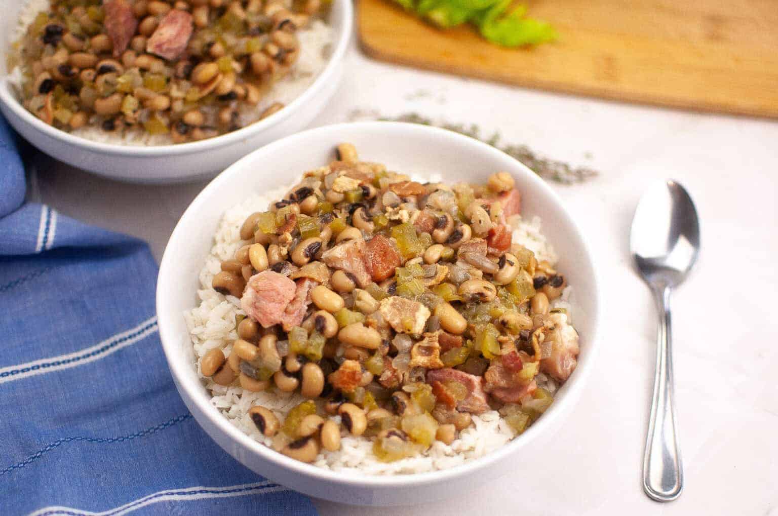 A bowl of Hoppin’ John served over white rice, with a spoon and a blue napkin nearby.