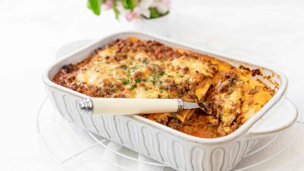 A baked lasagna in a white ceramic dish with a serving spatula, showing layers of pasta, meat sauce, and melted cheese.