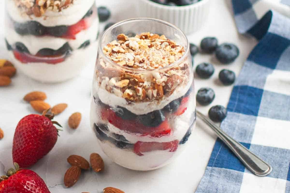 A glass filled with layers of cream, strawberries, blueberries, and almond nuts sits on a table with whole strawberries, blueberries, almonds, and a spoon nearby.