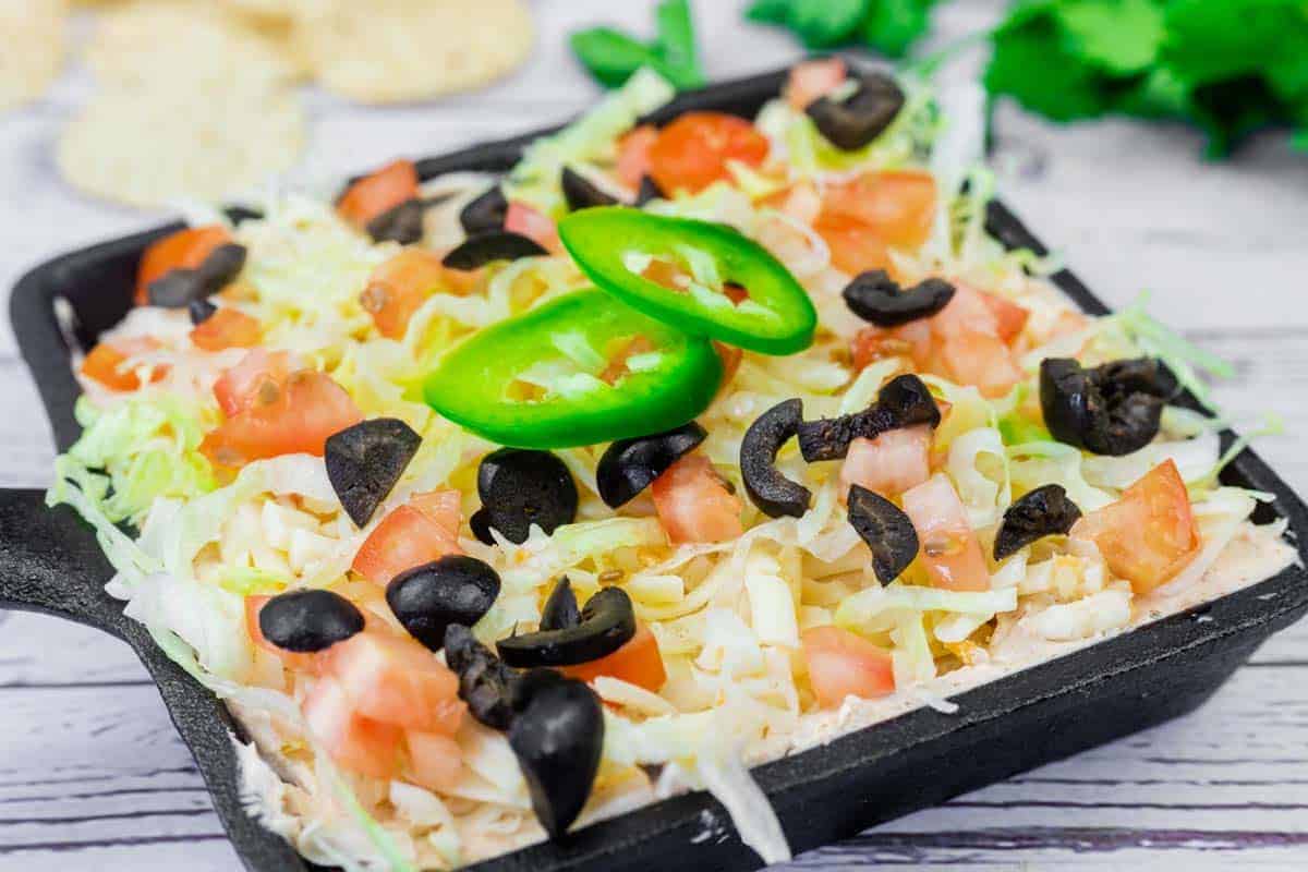 A skillet of Taco dip filled with shredded lettuce, diced tomatoes, sliced black olives, and jalapeño slices, served on a light wooden surface.