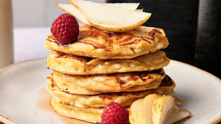 A stack of pancakes topped with sliced pear and raspberries, served on a beige plate with extra pear slices and raspberries on the side.