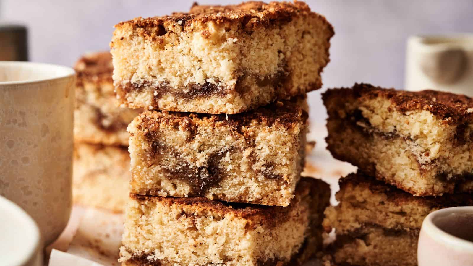 Three pieces of crumbly coffee cake with a cinnamon swirl are stacked on a plate, with two white mugs partially visible around them.