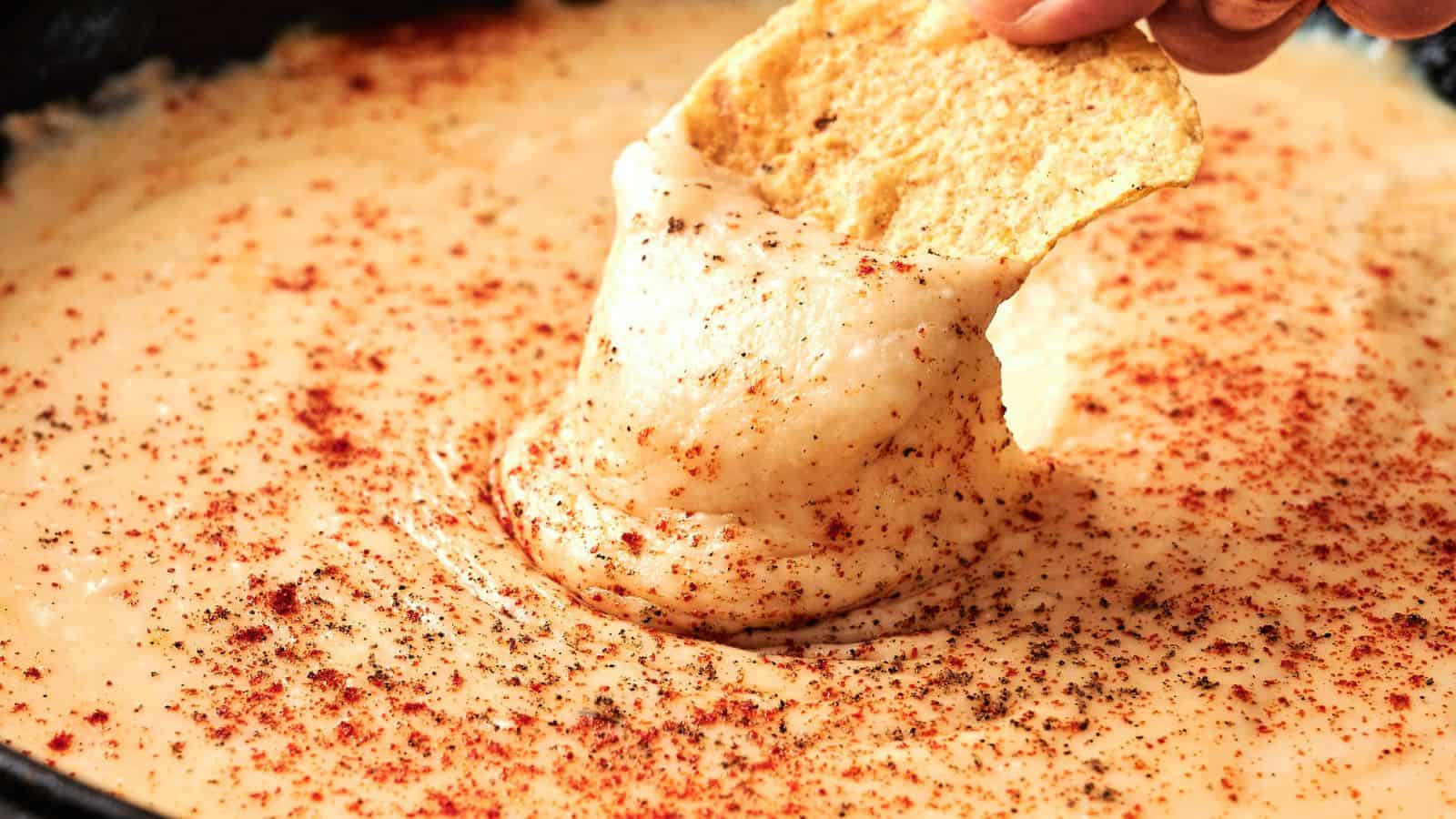 A hand dips a tortilla chip into creamy cheese dip sprinkled with paprika and black pepper.