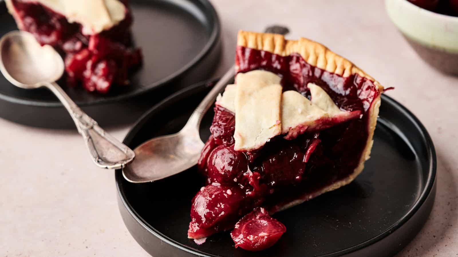 A slice of cherry pie with a flaky crust is served on a black plate with a spoon; another plate with pie is in the background.